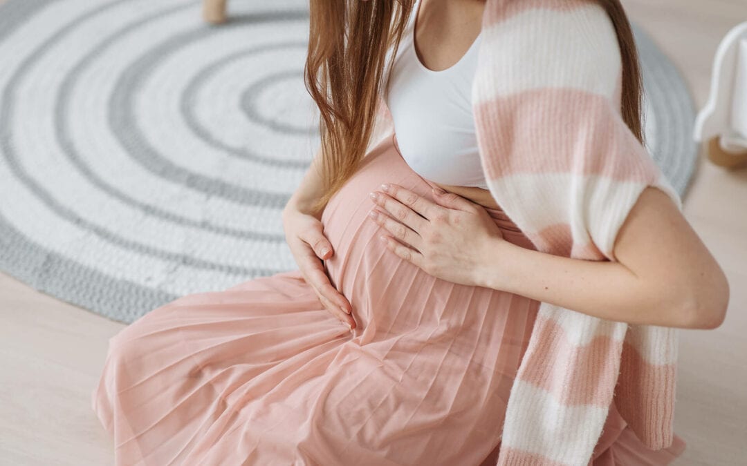 A woman in a white top and pink skirt, draped in a striped shawl, sits on the floor holding her pregnant belly with both hands.