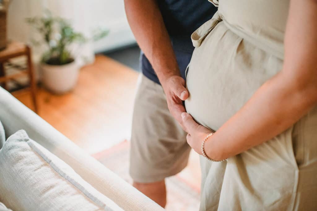 A woman in a beige dress cradles her pregnant belly while a man in a dark shirt and shorts stands beside her, touching her belly.