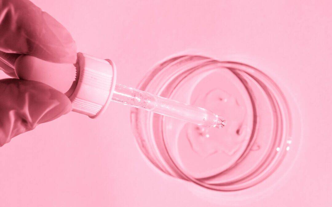 Gloved hand holding a dropper above a petri dish containing a liquid substance. Image in pink hue.