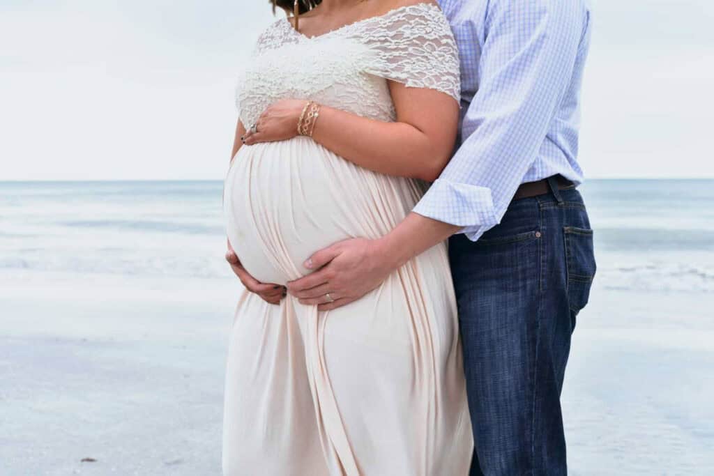 A pregnant woman in a white dress stands on a beach, with a man in a striped shirt embracing her and holding her belly from behind.