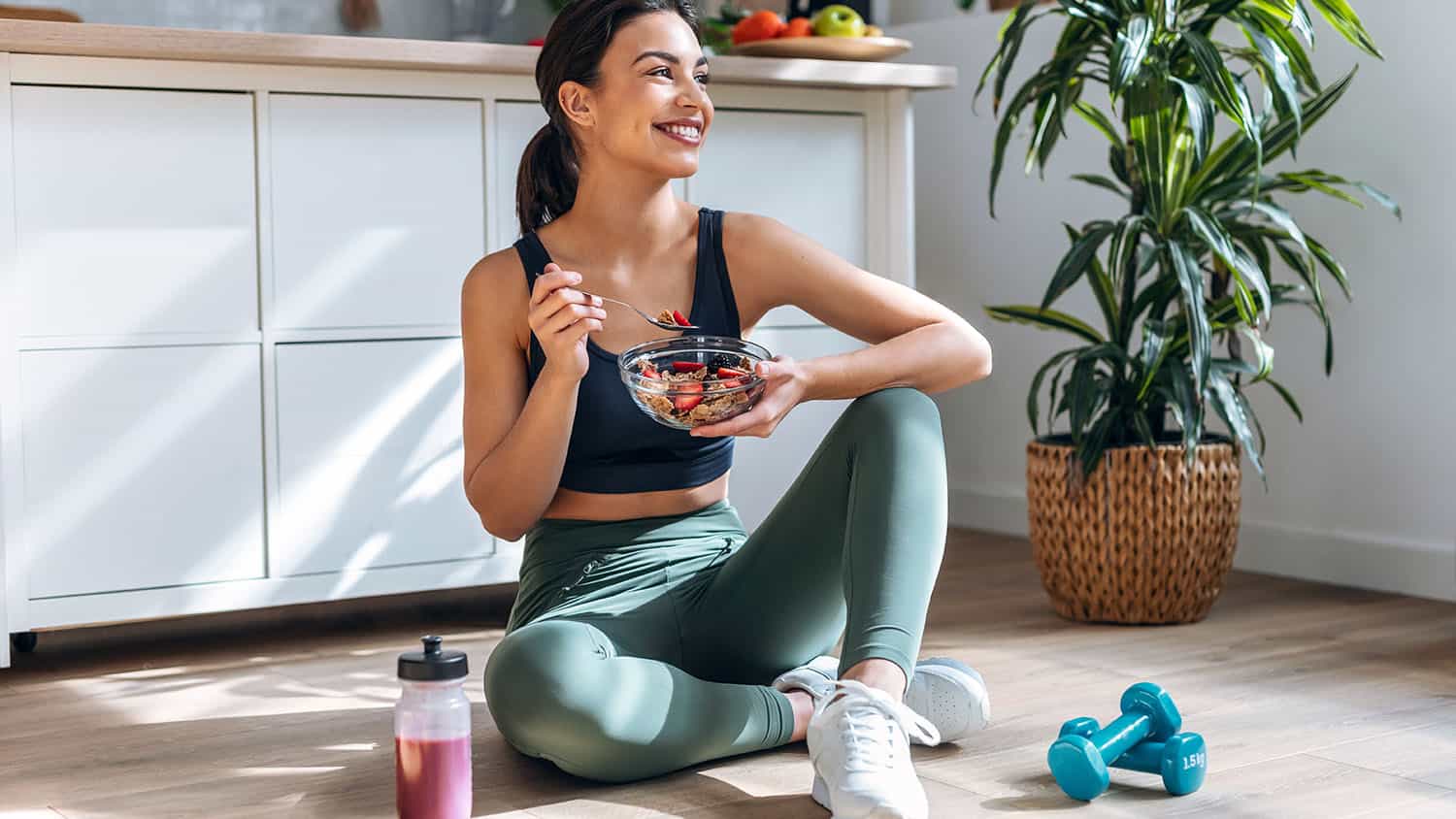 Shot of athletic woman eating a healthy bowl of muesli with fruit sitting on floor in the kitchen at home